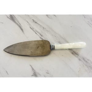 May include: A vintage cake server with a stainless steel blade and a white, pearlescent handle. The blade has a serrated edge and a slightly tarnished appearance. The handle is attached with silver-colored metal accents. The server is designed for cutting and serving cake.