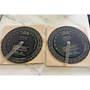May include: Two circular black aviation flight computers, A.C. Type G-1, mounted on square cardboard bases. Each device features a rotating dial with white printed scales, text, and a clear plastic indicator arm for calculating true airspeed.