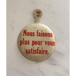 May include: A vintage, off-white button with a red inscription in French: "Nous faisons plus pour vous satisfaire." The button has a small, attached handle with a faded emblem. The overall design is circular.
