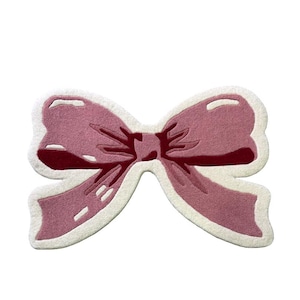 May include: A pink bow-shaped rug with a white border. The rug features a detailed design with varying shades of pink, creating a three-dimensional effect. The bow's center is a darker shade of pink, adding depth to the design.