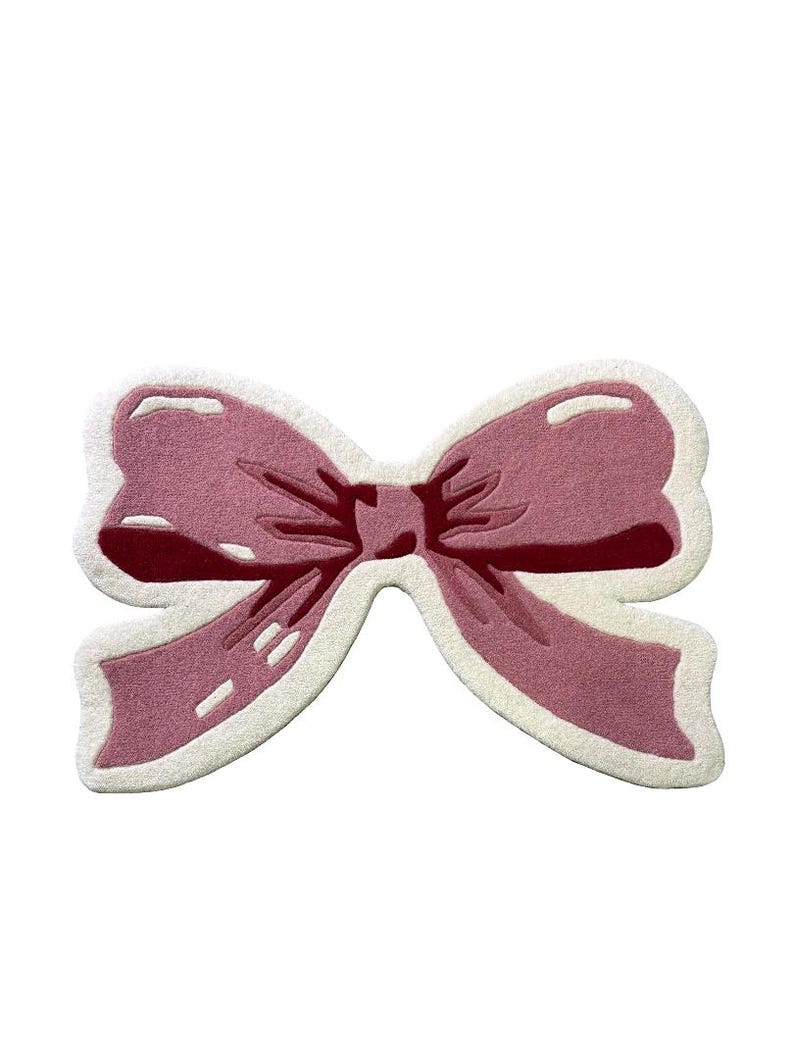 May include: A pink bow-shaped rug with a white border. The rug features a detailed design with varying shades of pink, creating a three-dimensional effect. The bow's center is a darker shade of pink, adding depth to the design.