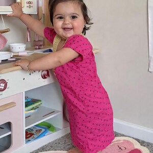 May include: A young child in a pink polka-dot dress smiles while playing with a wooden toy kitchen. The kitchen has a microwave, stove, and shelves with toy accessories. A pink and white butterfly rug is on the floor.