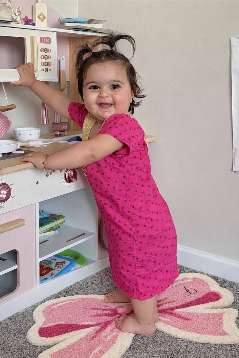 May include: A young child in a pink polka-dot dress smiles while playing with a wooden toy kitchen. The kitchen has a microwave, stove, and shelves with toy accessories. A pink and white butterfly rug is on the floor.