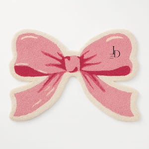 May include: A pink bow-shaped rug with a cream-colored border. The bow has a textured appearance and a darker pink center. The rug features a logo in the upper right corner.