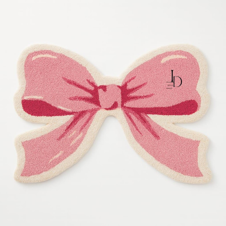 May include: A pink bow-shaped rug with a cream-colored border. The bow has a textured appearance and a darker pink center. The rug features a logo in the upper right corner.