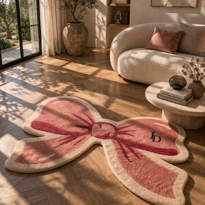 May include: A pink and beige bow-shaped rug with a textured surface. The rug features a darker pink outline and a central knot design. The initials "JD" are embroidered on one of the bow loops. The rug is placed on a wooden floor.