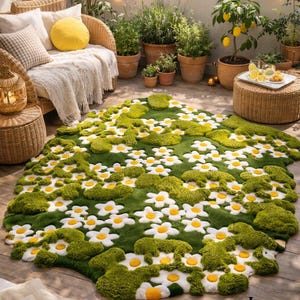 May include: A green rug with a daisy flower design. The rug features white daisy petals with yellow centers, set against a green background. The rug is placed on a wooden deck, with outdoor furniture and plants in the background.