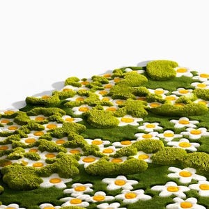May include: A green rug with a daisy flower design. The rug features white daisy petals with yellow centers, surrounded by green moss-like tufts. The rug has a soft, textured appearance, creating a whimsical, natural aesthetic. The logo "JD RUGS" is in the top left corner.