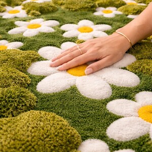 May include: A daisy-shaped rug with white petals, yellow centers, and green grass-like background. The rug has a soft, textured appearance. A hand with a gold ring and bracelet rests on the rug.