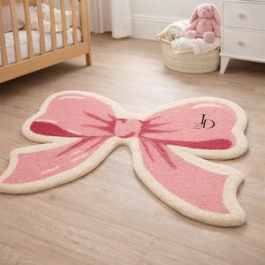 May include: A pink bow-shaped rug with a cream-colored border. The rug has a textured surface and a darker pink center. The initials "JD" are printed on the right side of the bow. The rug is on a light wood floor.