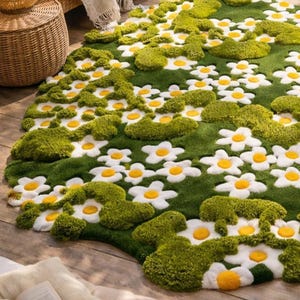 May include: A green rug with a daisy flower design. The rug features white daisy flowers with yellow centers and green moss-like accents. The rug is placed on a wooden floor, creating a natural, floral theme.