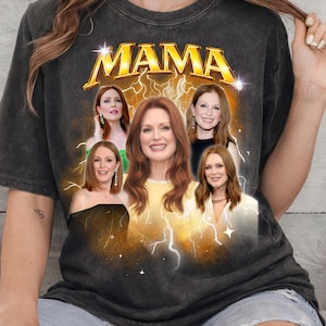May include: Dark gray t-shirt featuring the word "MAMA" in gold lettering above a collage of images. The images are of a woman with red hair, set against a background of lightning bolts and stars.