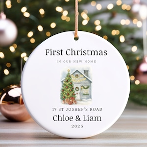 Może przedstawiać: Biała, okrągła ozdoba świąteczna z napisem "First Christmas In Our New Home". Zawiera akwarelową ilustrację domu z choinką i prezentami. Ozdoba zawiera adres, imiona i rok 2025.