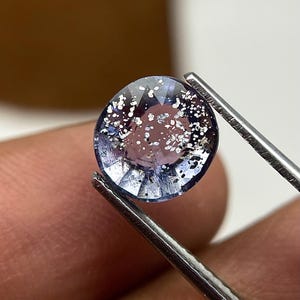 May include: A round, light blue gemstone with internal sparkle, held by tweezers. The gem's facets are visible, reflecting light. The inclusions within the stone add to its visual appeal. The tweezers provide a close-up view of the gem's details.