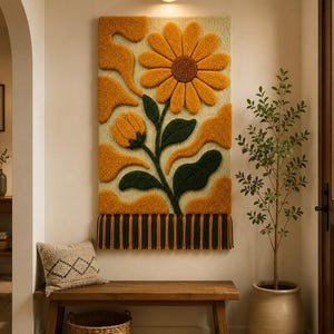 May include: A rectangular tufted wall hanging featuring a large yellow sunflower and a smaller bud with dark green leaves against a cream background. The design includes abstract orange shapes and a bottom fringe of alternating green and orange yarn.