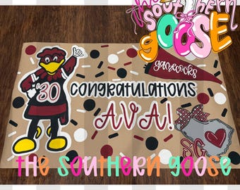 May include: A brown paper banner with the text "Congratulations Ava!" and a cartoon goose wearing a pink hat. The banner also features a cartoon mascot, confetti, and the words "Gamecocks". The Southern Goose logo is also visible.