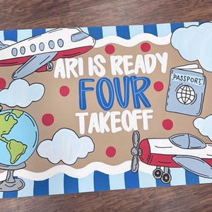 May include: A rectangular sign with a light blue and dark blue striped border featuring a tan center with red dots. It displays the text ARI IS READY FOUR TAKEOFF, a white airplane, a blue globe, a light blue passport, and white clouds.
