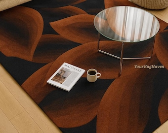 May include: A modern area rug with a black background and large, abstract brown leaf-like shapes. A glass-topped coffee table, a beige sofa, and a woven ottoman are in the background. A magazine and a coffee cup sit on the rug.