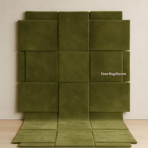 May include: Olive green woven rug with a geometric design. The rug is made of rectangular panels, some of which are raised, creating a textured surface. The rug is displayed against a neutral wall and wooden floor.