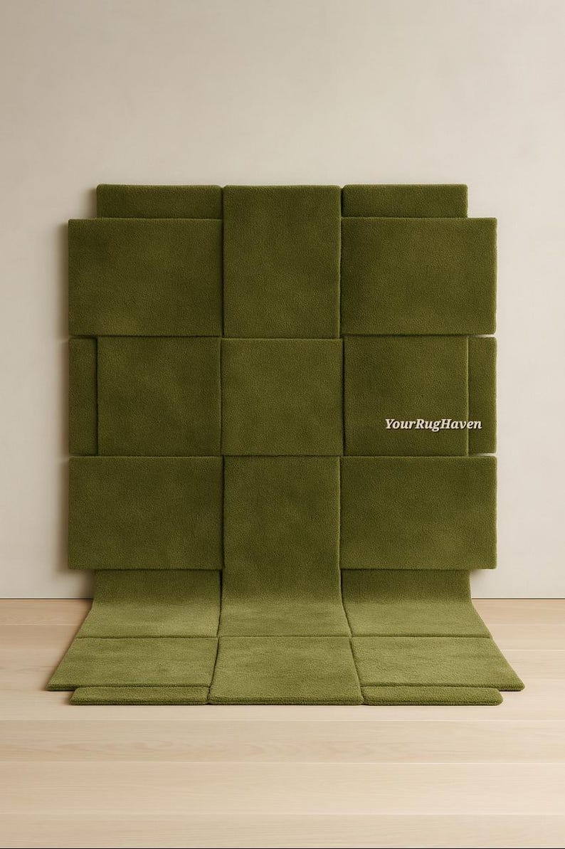 May include: Olive green woven rug with a geometric design. The rug is made of rectangular panels, some of which are raised, creating a textured surface. The rug is displayed against a neutral wall and wooden floor.