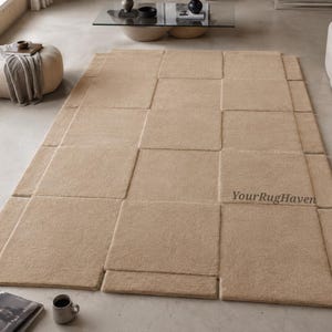 May include: A rectangular area rug in a solid beige color featuring a geometric pattern of overlapping square and rectangular segments. The rug has a textured, plush surface and includes the text YourRugHaven printed on the lower right side.