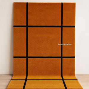 May include: Mustard-colored rectangular rug with a black grid pattern. The rug is made of a soft material and has a modern design. The text "YourRugHaven" is visible on the rug.