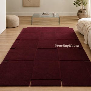 May include: A rectangular burgundy rug with a geometric design, composed of multiple square and rectangular sections. The rug is placed on a light-colored wooden floor. The text "YourRugHaven" is visible on the rug.
