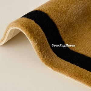 May include: Close-up of a rolled-up rug with a tan background and a thick black stripe. The rug has a soft, plush texture. The text "YourRugHaven" is visible on the rug.