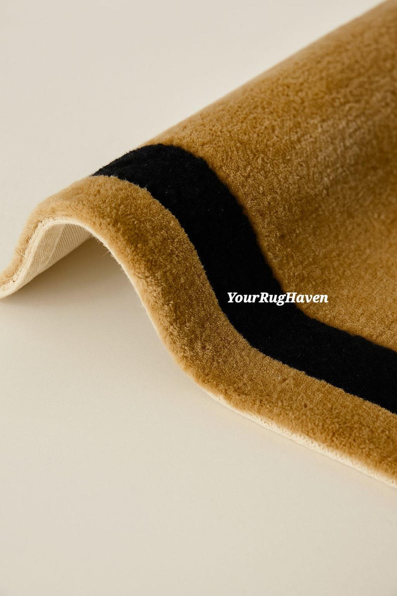 May include: Close-up of a rolled-up rug with a tan background and a thick black stripe. The rug has a soft, plush texture. The text "YourRugHaven" is visible on the rug.