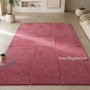 May include: A rectangular area rug in a deep dusty rose color, featuring a geometric grid pattern composed of multiple square and rectangular segments with visible seams. The text YourRugHaven is printed in white on the right side of the rug.