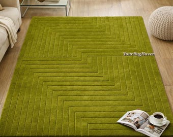 May include: A large, rectangular green rug with a geometric pattern of angled lines. The rug is the focal point of a living room setting, with a coffee table, sofa, and ottoman visible. A magazine and a cup of coffee sit on the rug.