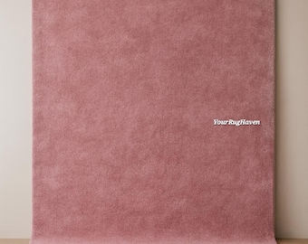 Puede incluir: Una alfombra rectangular de color rosa polvoriento sólido con una textura densa de pelo corto. La alfombra está colocada verticalmente contra una pared y horizontalmente en el suelo. El texto YourRugHaven está impreso en blanco a la derecha.