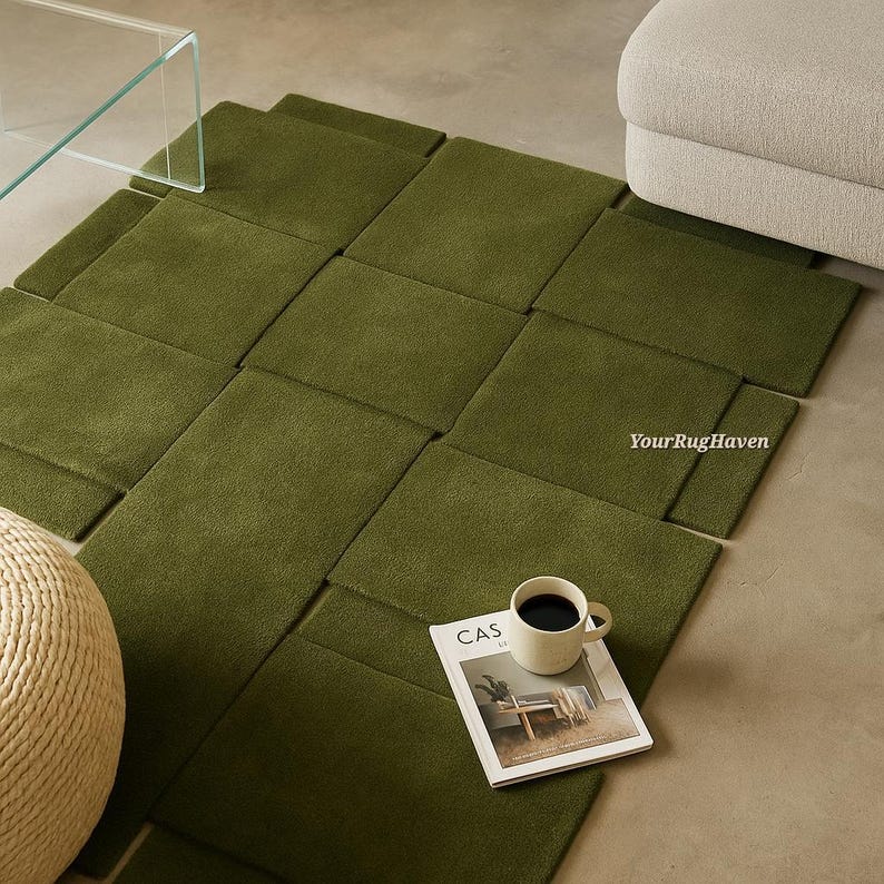 May include: An olive green woven area rug with a geometric design. A coffee cup and a magazine are on the rug. The magazine has the word "CAS" on the cover. The rug is near a beige ottoman and a glass table.