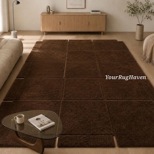 May include: A large, modular brown rug composed of rectangular and square sections, arranged on a wooden floor. A glass coffee table with a book and mug sits in front of the rug. The rug covers a large area of the room.