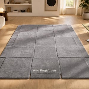 May include: A large, rectangular gray rug with a geometric design. The rug is composed of multiple square and rectangular sections, creating a modern, modular look. The rug is placed on a hardwood floor in a living room setting.