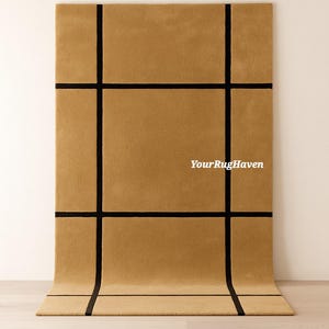 May include: A rectangular, tan-colored rug with a grid pattern of thick, black lines. The rug is displayed on a light wood floor, with the top portion against a white wall. The text "YourRugHaven" is visible on the rug.