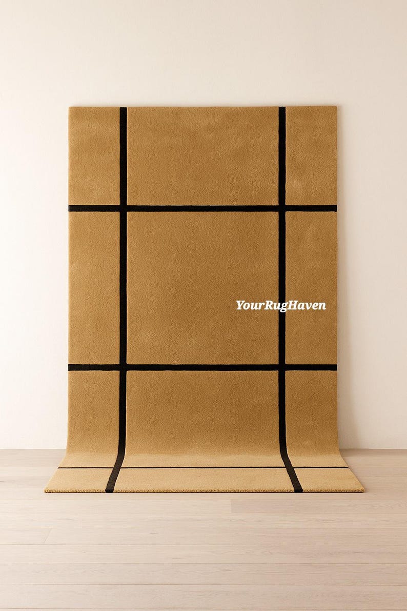 May include: A rectangular, tan-colored rug with a grid pattern of thick, black lines. The rug is displayed on a light wood floor, with the top portion against a white wall. The text "YourRugHaven" is visible on the rug.