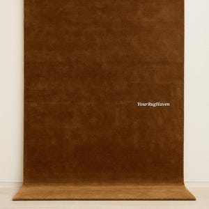 May include: A rectangular area rug in a solid, deep brown color with a soft, textured pile surface. The text "YourRugHaven" is printed in white on the right side of the rug. The rug is positioned vertically against a plain, light-colored wall.