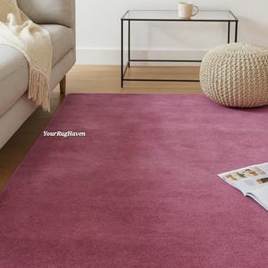 May include: A large, rectangular rug in a deep rose colour fills the foreground. The rug is in a living room setting, with a light grey sofa, a black side table, and a woven ottoman in the background. The text "YourRugHaven" is visible on the rug.