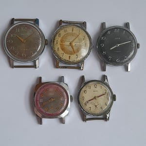 Pode incluir: Uma coleção de cinco relógios de pulso vintage com caixas de metal prateado. Os relógios apresentam mostradores redondos em várias cores, incluindo castanho, creme, preto e rosa, com algarismos arábicos, ponteiros finos e coroas laterais.