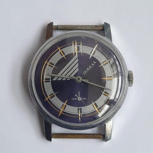 May include: A vintage wristwatch featuring a dark blue face with silver and gold-tone detailing. The watch face displays the Cyrillic word "ПОБЕДА". The case is silver-tone, and the watch has a textured crown. The hands are gold-tone, with a classic design.
