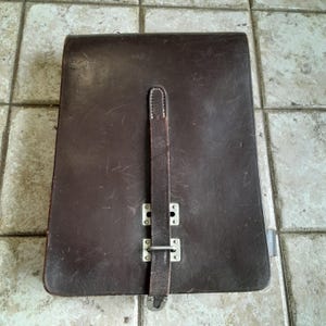 Puede incluir: Un maletín de cuero marrón oscuro con una correa vertical y cierre de hebilla metálica. La funda rectangular tiene un aspecto ligeramente desgastado, lo que sugiere edad y uso. La correa está asegurada con herrajes metálicos.