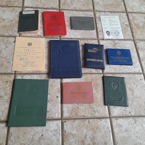 May include: A collection of various vintage documents and identification booklets in different colors including red, blue, green, and pink. The set features hard-cover folders, paper certificates with Cyrillic text, and small rectangular cards with emblems.