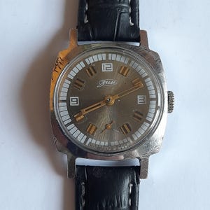 Könnte beinhalten: Vintage silberfarbene Armbanduhr mit grauem Zifferblatt, goldenen Zeigern und schwarzem Lederarmband. Das Zifferblatt hat weiße rechteckige Stundenmarkierungen und die Zahlen 12, 3 und 9. Die Uhr trägt den Text "Сделано в СССР".