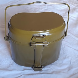 May include: Olive green metal mess kit with a handle and a hinged lid. The kit has a rounded rectangular shape with a handle for carrying. The lid is secured with a metal clasp. The mess kit is designed for outdoor use.