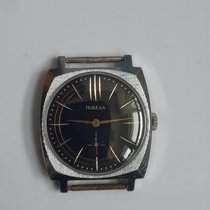 Puede incluir: Reloj de pulsera vintage plateado con esfera negra y agujas e indicadores dorados. La esfera del reloj tiene la palabra "ПОБЕДА" en letras doradas. El reloj tiene una caja rectangular y un bisel texturizado.