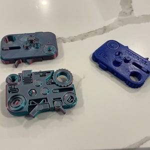 May include: Three 3D-printed game controller designs in blue and gray. One blue controller has the text "MIXITURE" on it. The other two controllers are gray with red and blue accents. The controllers have buttons, joysticks, and other details.