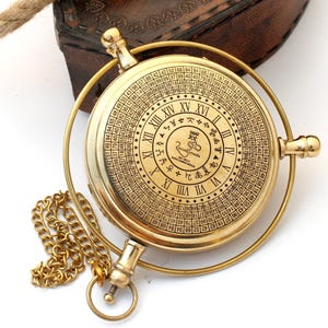 May include: A vintage brass astrolabe with a gold chain and a leather case in the background. The astrolabe features Roman numerals and other symbols. The astrolabe is a navigational instrument.
