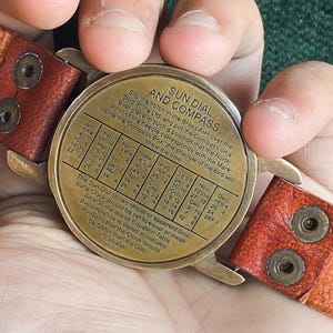May include: A vintage brass sun dial and compass with a leather strap. The dial features text and a table for time conversion. The leather strap is brown with visible stitching and metal rivets. The dial is held in a person's hands.