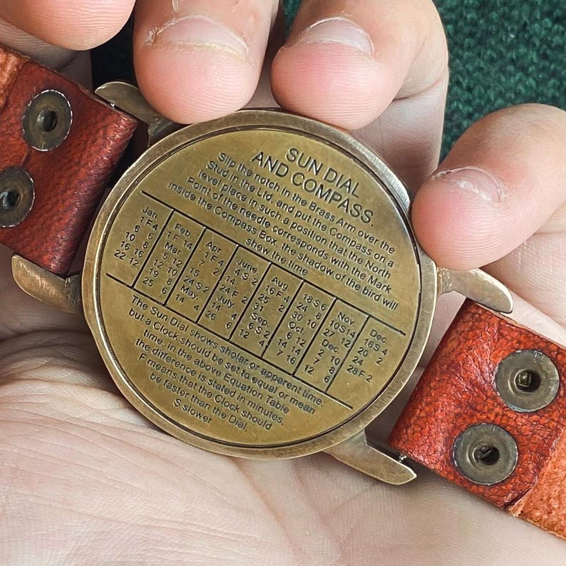 May include: A vintage brass sun dial and compass with a leather strap. The dial features text and a table for time conversion. The leather strap is brown with visible stitching and metal rivets. The dial is held in a person's hands.